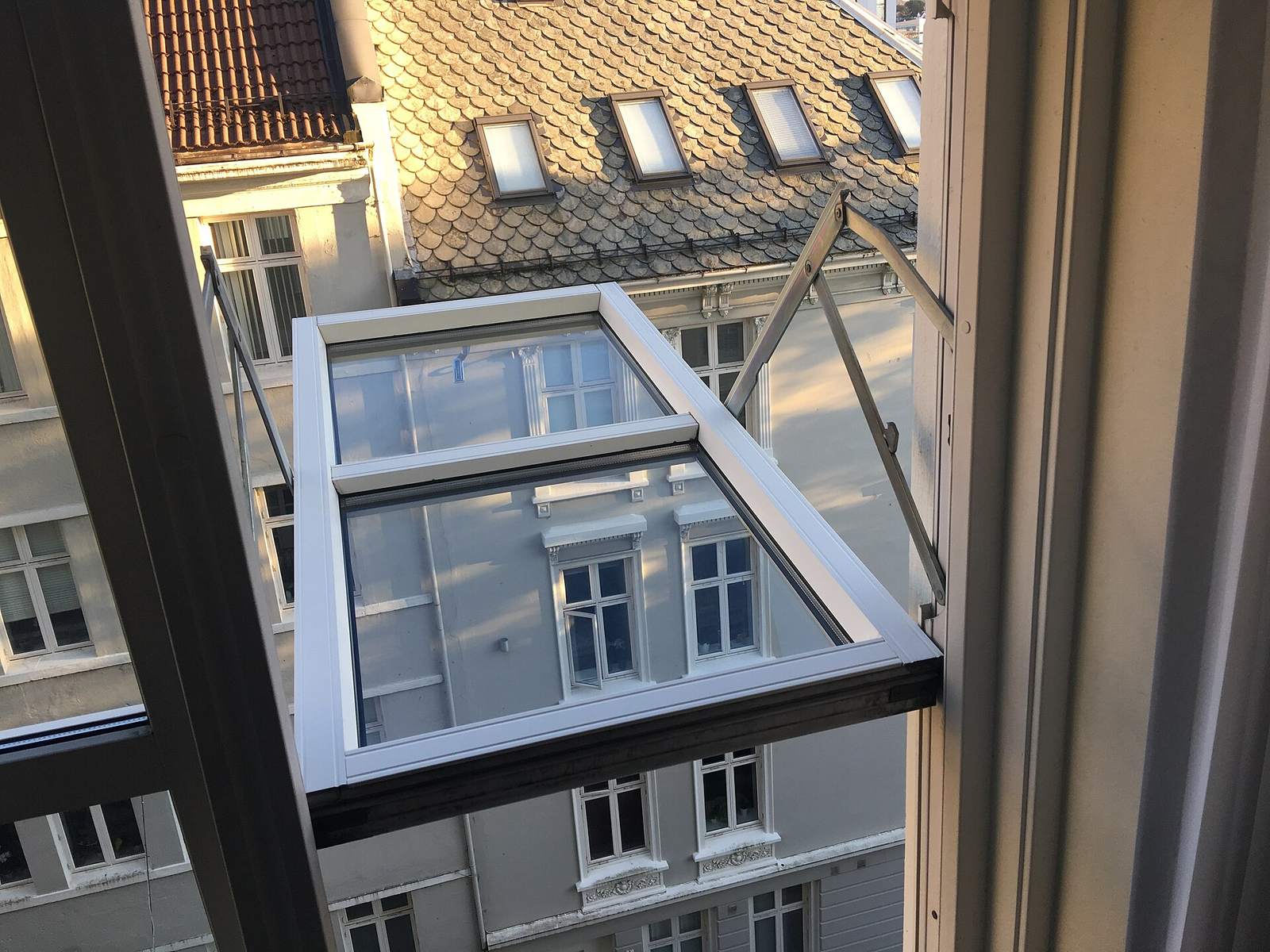Wijd opengezet raam hoog in een stadsgebouw, met zicht op daken met dakramen en witte gevels ertegenover